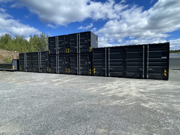 Svarte miljøcontainere stablet i to høyder på Nordcons lagerområde. Bildet er tatt utendørs med blå himmel og skyer i bakgrunnen.